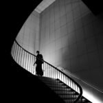 A silhouette of a person ascending a modern, curved staircase in black and white.